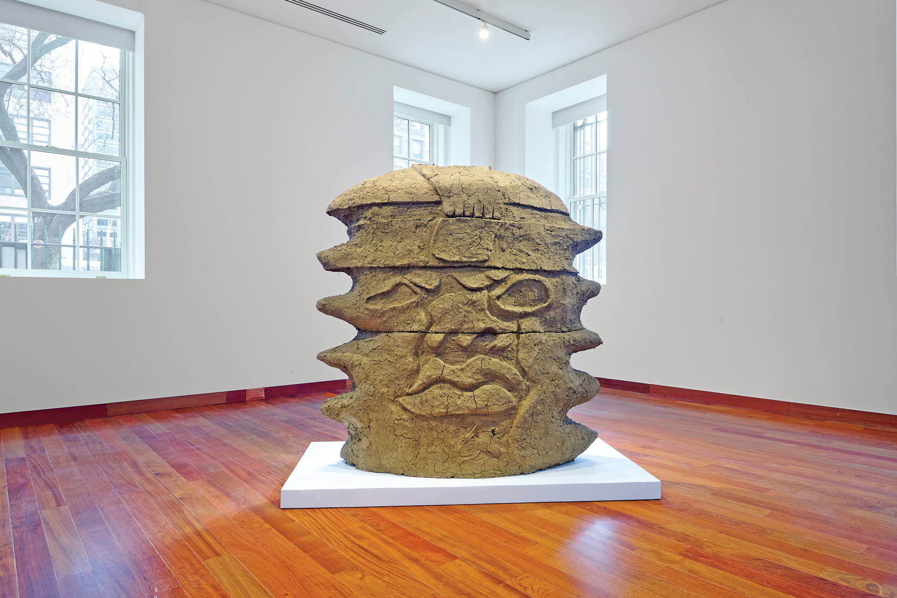 A large, textured clay sculpture with carved facial features, segmented and ridged, displayed on a white base in a gallery with wooden floors.