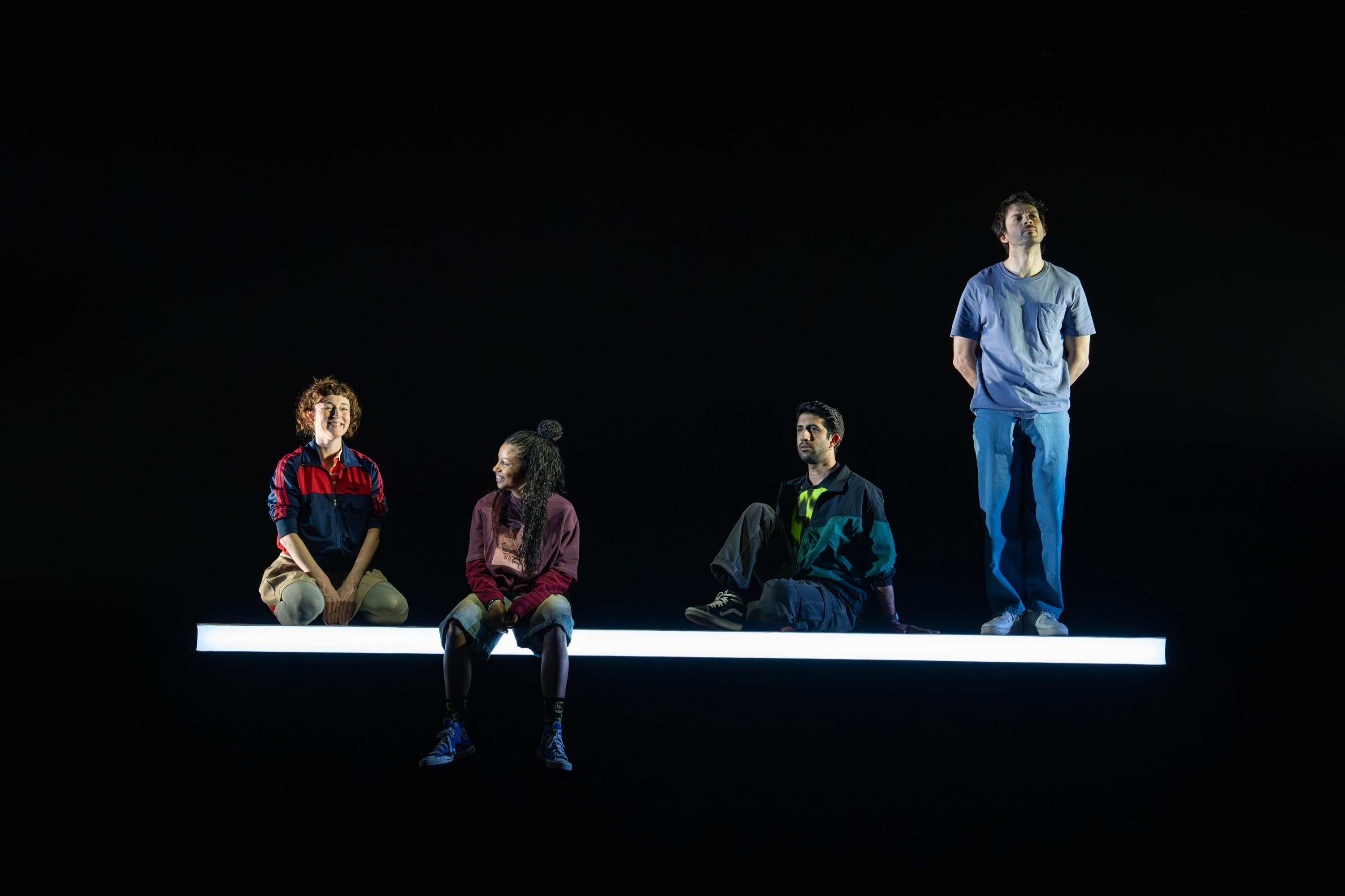 Four actors on a glowing white platform in a dark space, wearing casual clothing; two seated, one kneeling, and one standing.