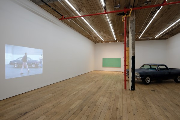 A gallery space with wooden floors, a video projection on a white wall, a green rectangular artwork, a vintage car, and red ceiling pipes with a pillar.