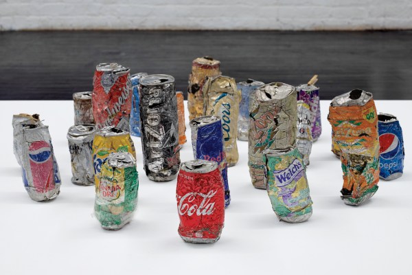 An installation featuring a group of crushed, colorful soda and beverage cans displayed on a white plinth, with brands like Coca-Cola, Pepsi, Arizona, and others visible.