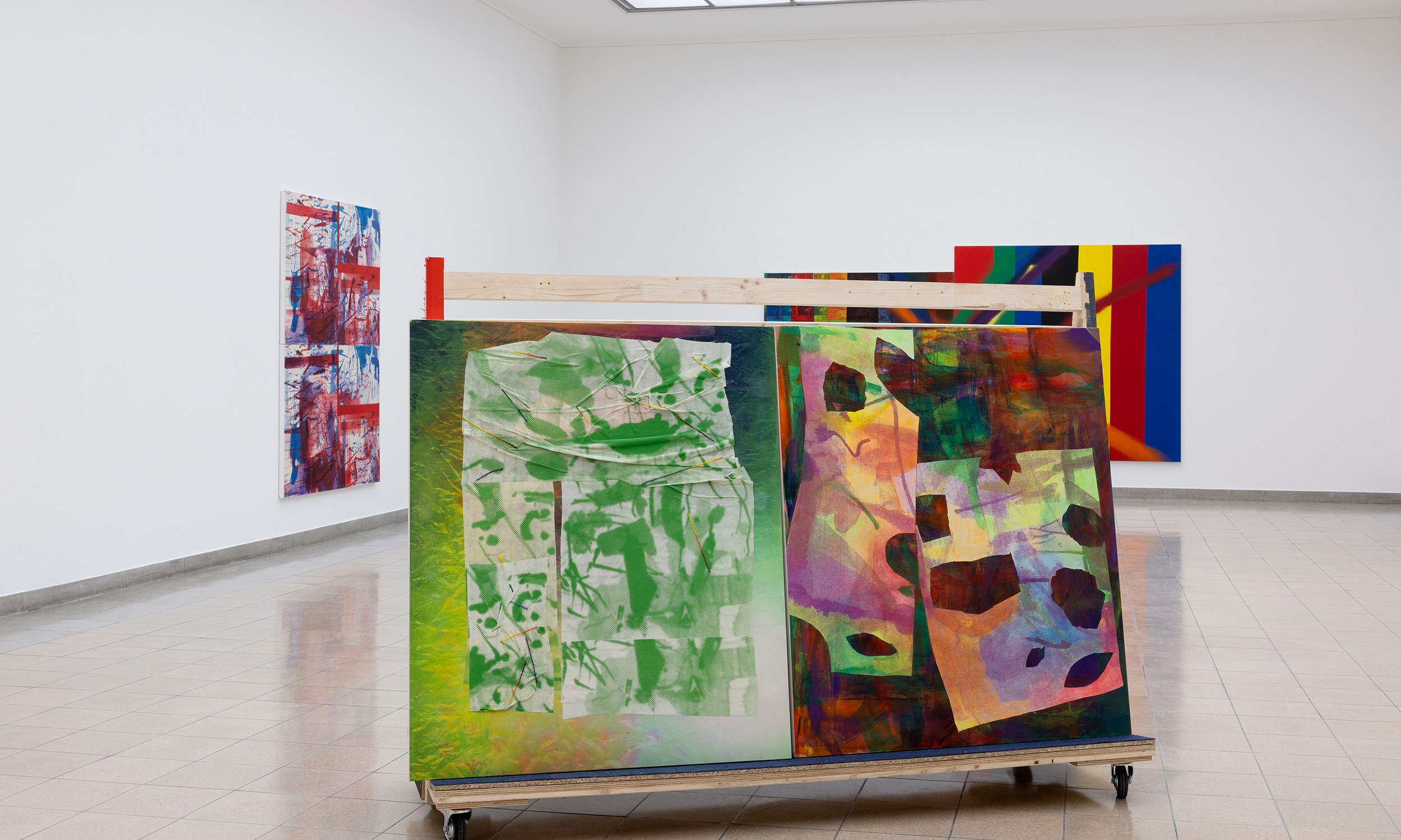 A gallery with abstract paintings; a two-sided artwork on a rolling display features green and multicolor designs, surrounded by wall-mounted pieces.