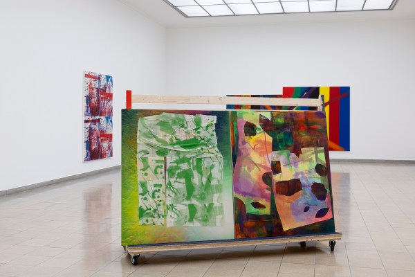 A gallery with abstract paintings; a two-sided artwork on a rolling display features green and multicolor designs, surrounded by wall-mounted pieces.
