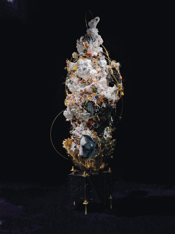 A vertical mixed-media sculpture with white porcelain shards, colorful debris, gold accents, wire loops, and intricate textures on a black pedestal.
