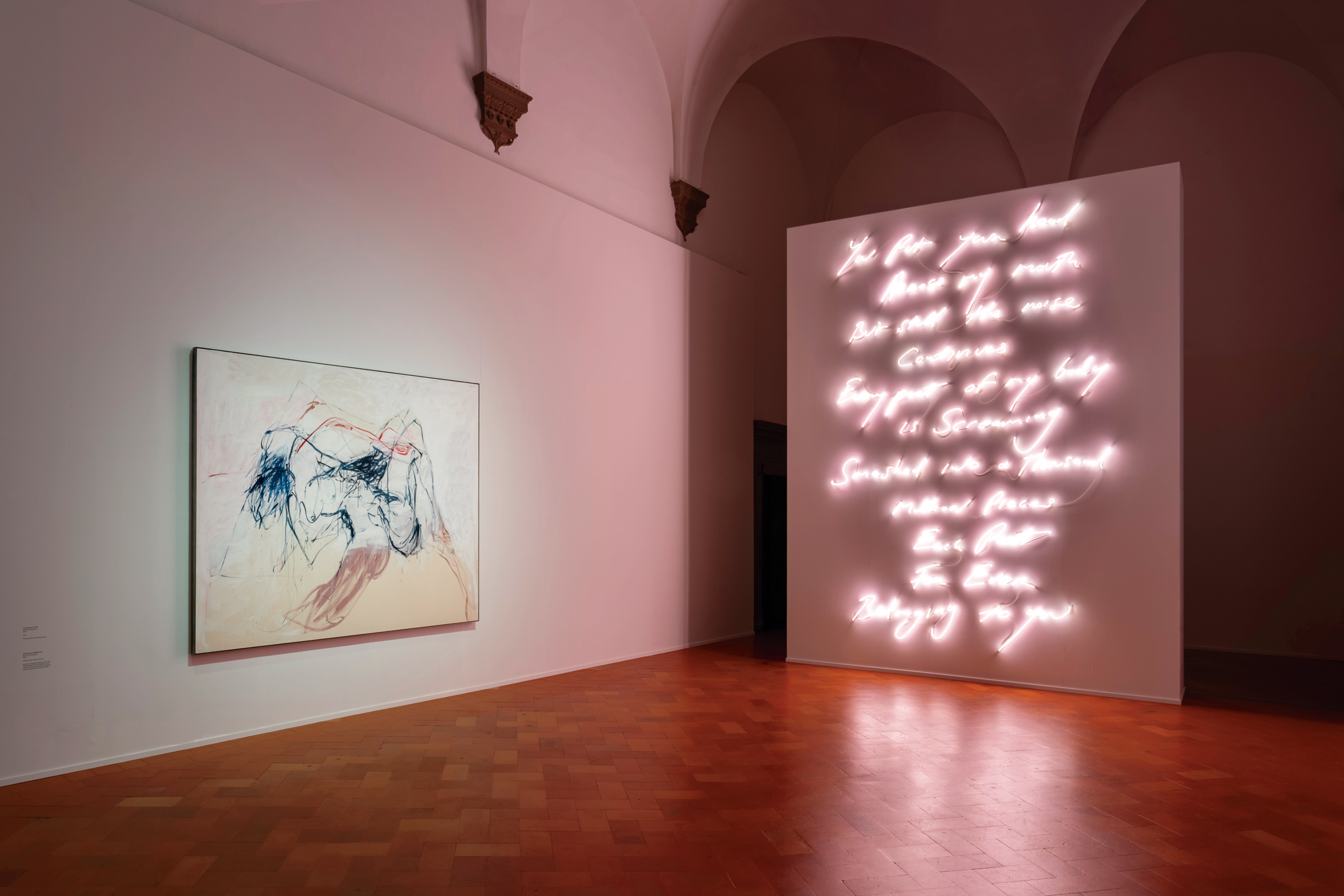 A room with vaulted ceilings, showcasing an abstract painting on the left and illuminated cursive neon text on the right, against white walls and tiled flooring.