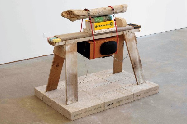 A sculpture composed of a wooden sawhorse with books, a speaker, a driftwood log, and various objects strapped on top, placed on a platform made of rectangular bricks.