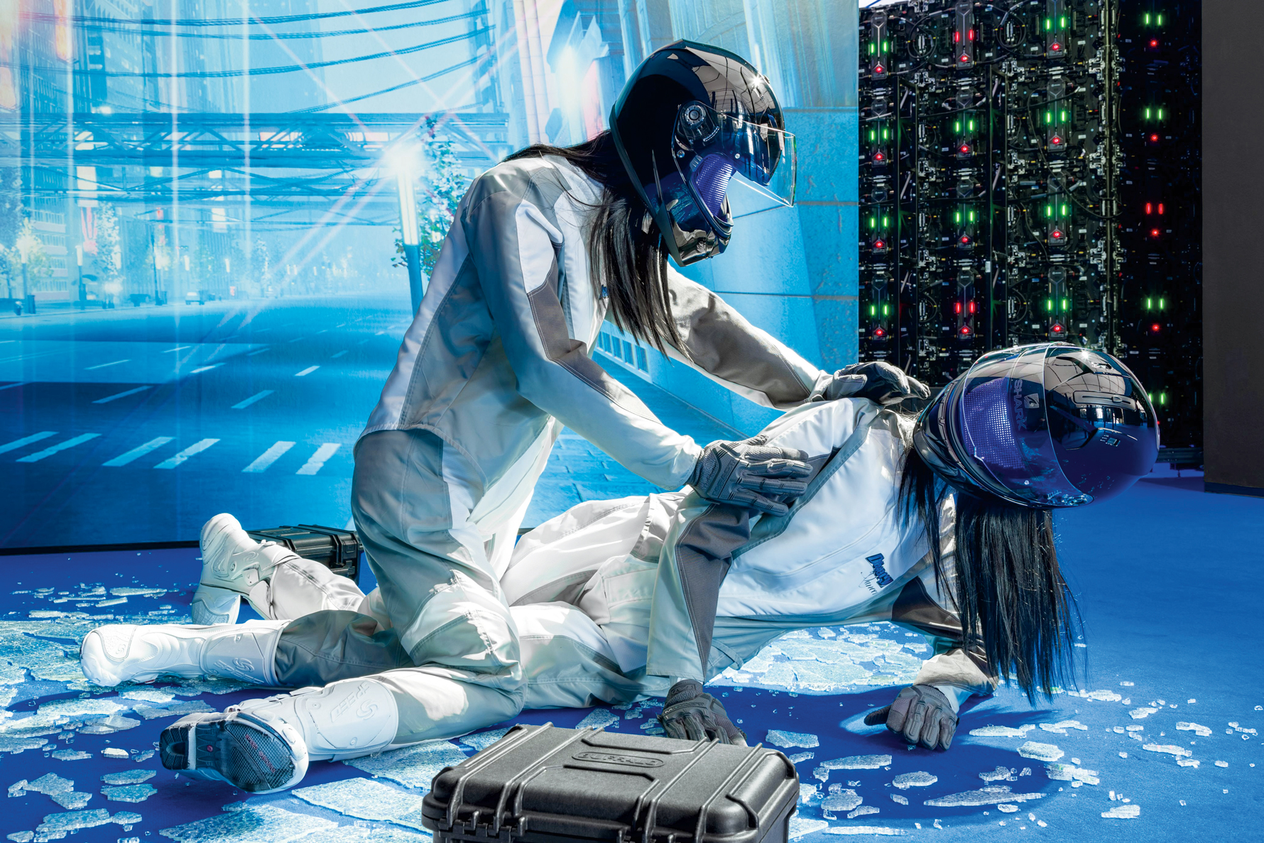 Two figures in helmets and futuristic white suits interact on a blue surface with scattered glass-like shards, a case nearby, and a digital urban backdrop.