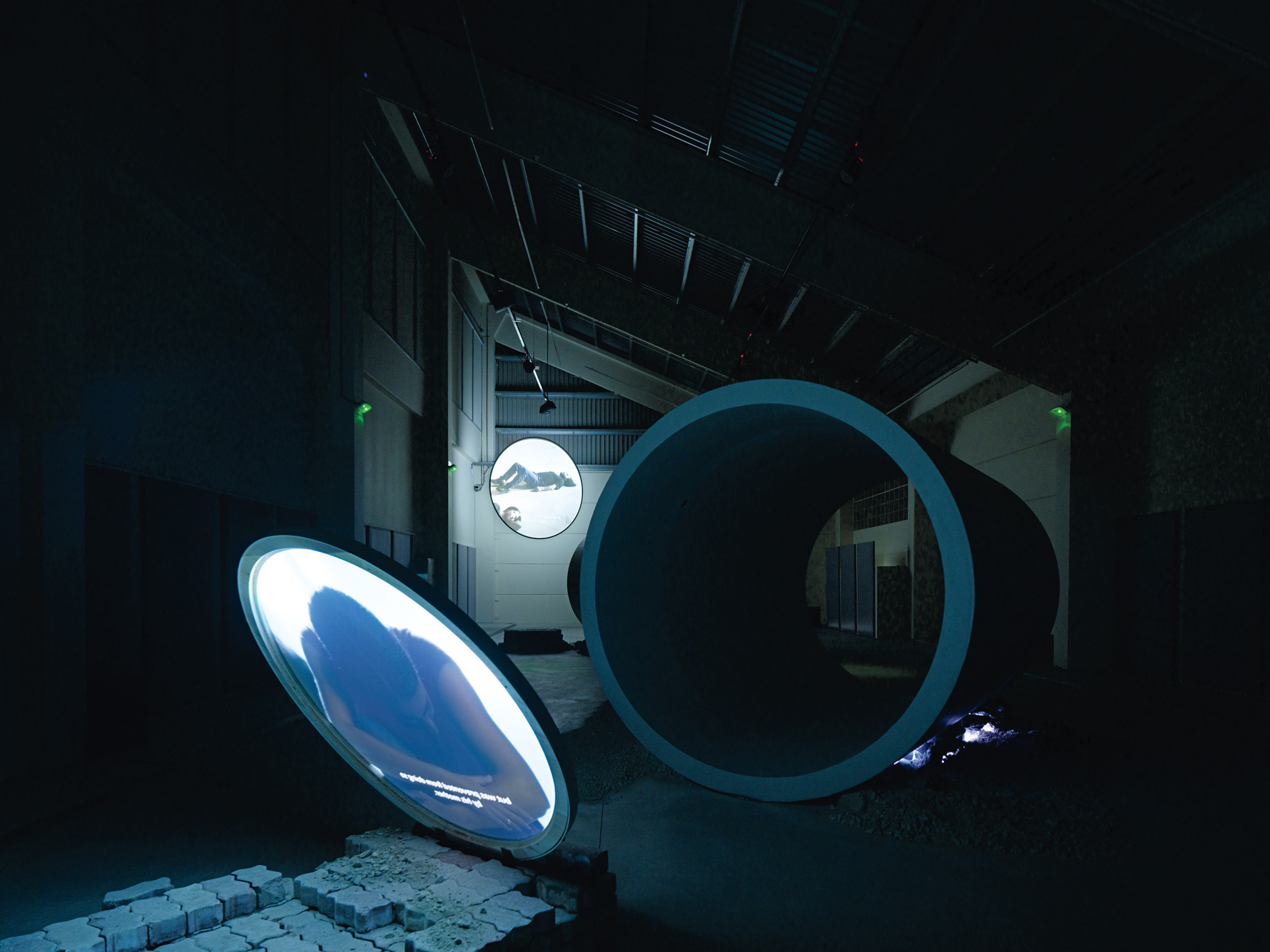 A dimly lit industrial space with large circular pipe structures, illuminated screens displaying imagery, and scattered bricks on the ground.
