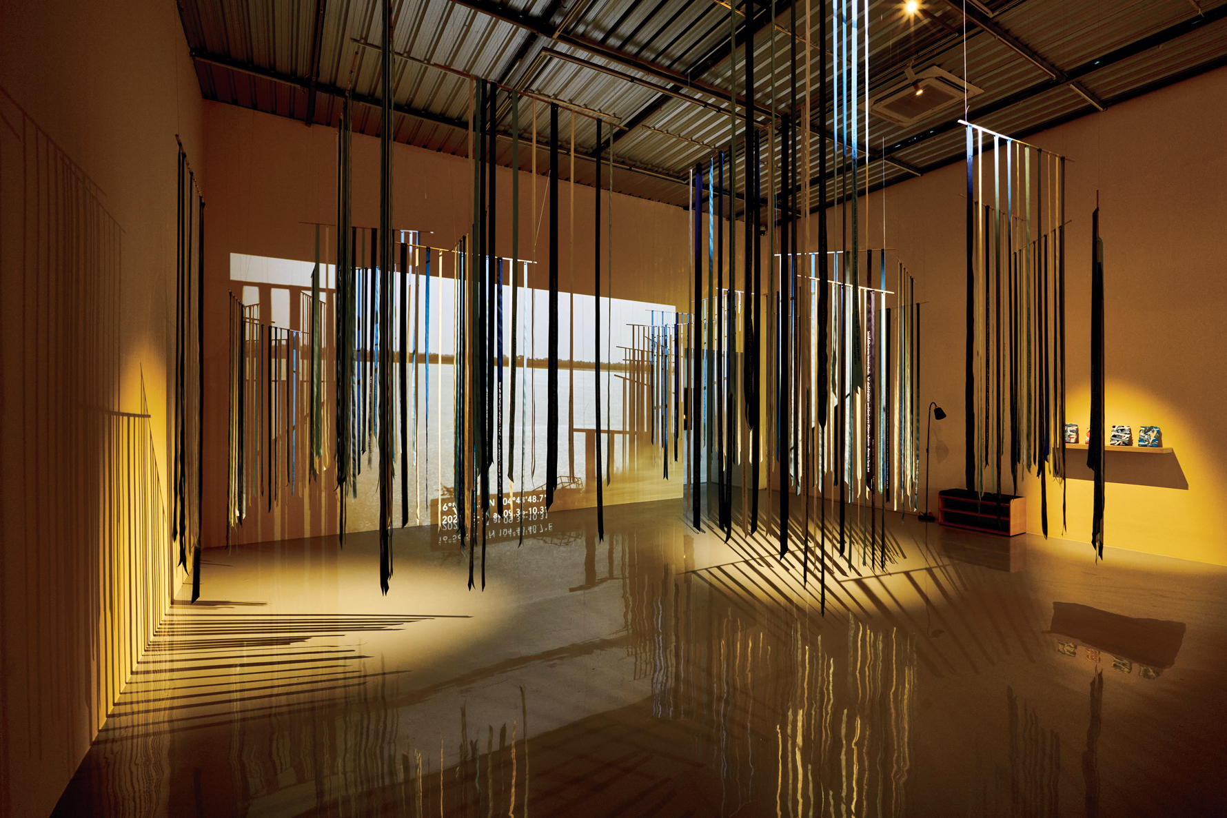 An installation with hanging vertical strips casting shadows, a reflective floor, a lamp, and shelves with items under warm lighting and projected visuals.