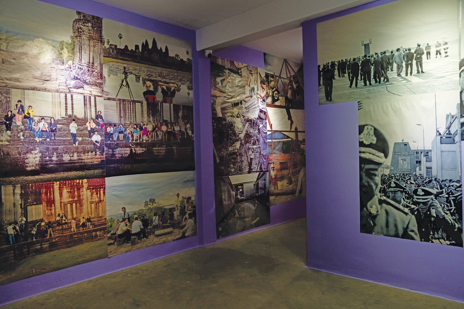 A gallery with purple walls displays large photographic prints depicting landscapes, people, historical sites, and black-and-white imagery of organized gatherings.