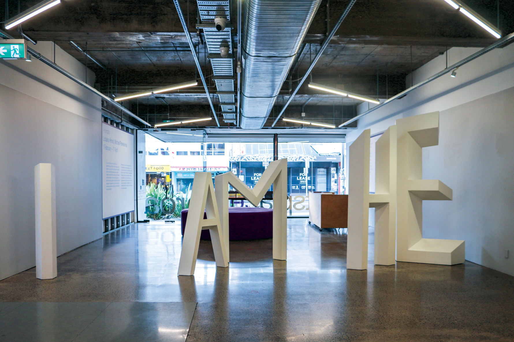 A museum space with large white 3D letters spelling "I AM HE," polished floors, exposed ceilings, and a glass storefront revealing an urban street.