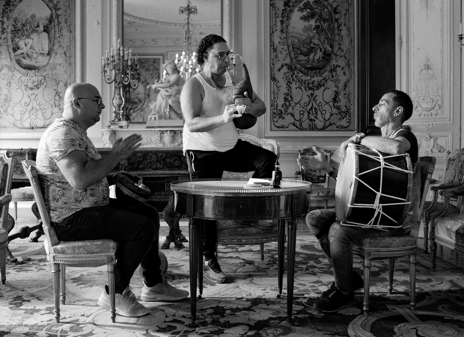 Three people are seated or standing in an ornate room with intricate wall art, chandeliers, a round table, and musical instruments like a drum and tambourine.