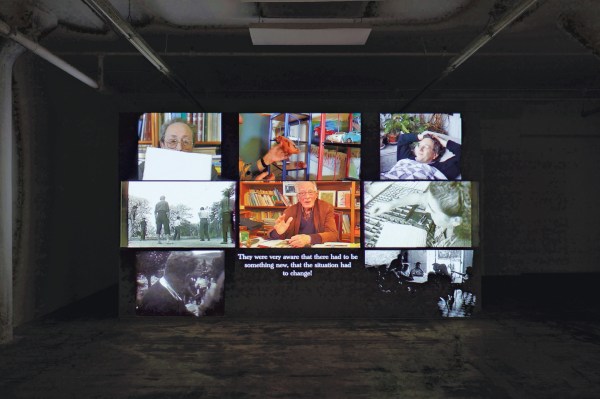 A wall display of video screens shows various scenes, including bookshelves, plants, people writing, outdoor activities, and text.