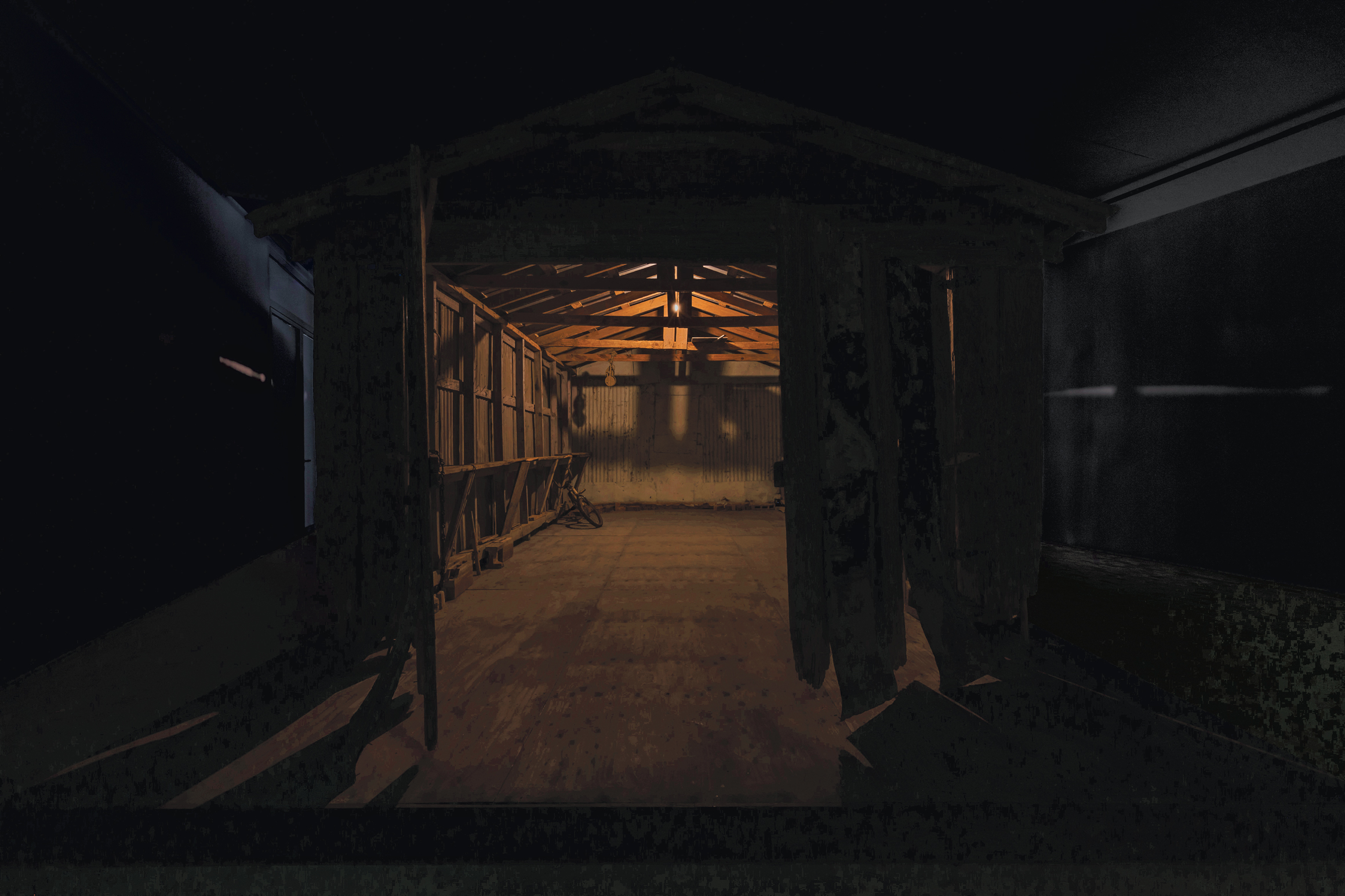 A dimly lit wooden structure with open doors, exposed beams, and a bicycle inside, surrounded by darkness with faint light on the walls.