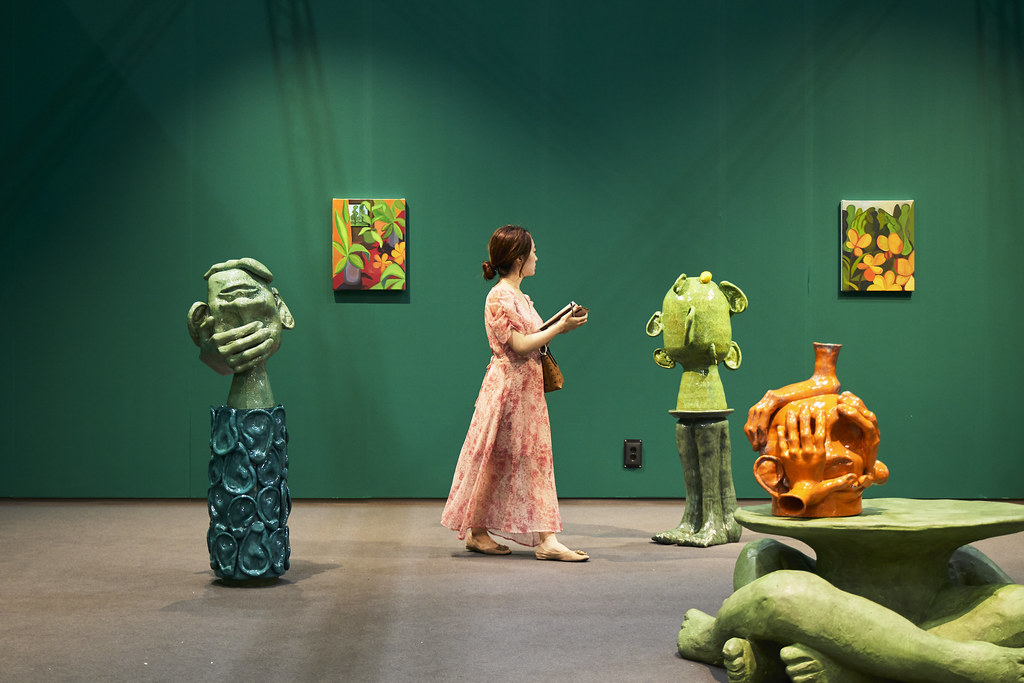 A woman in a pink dress exploring an art gallery. She is surrounded by colorful, abstract sculptures and two small paintings of flowers on a green wall. The sculptures are creative and unusual shapes, adding a playful feel to the space.