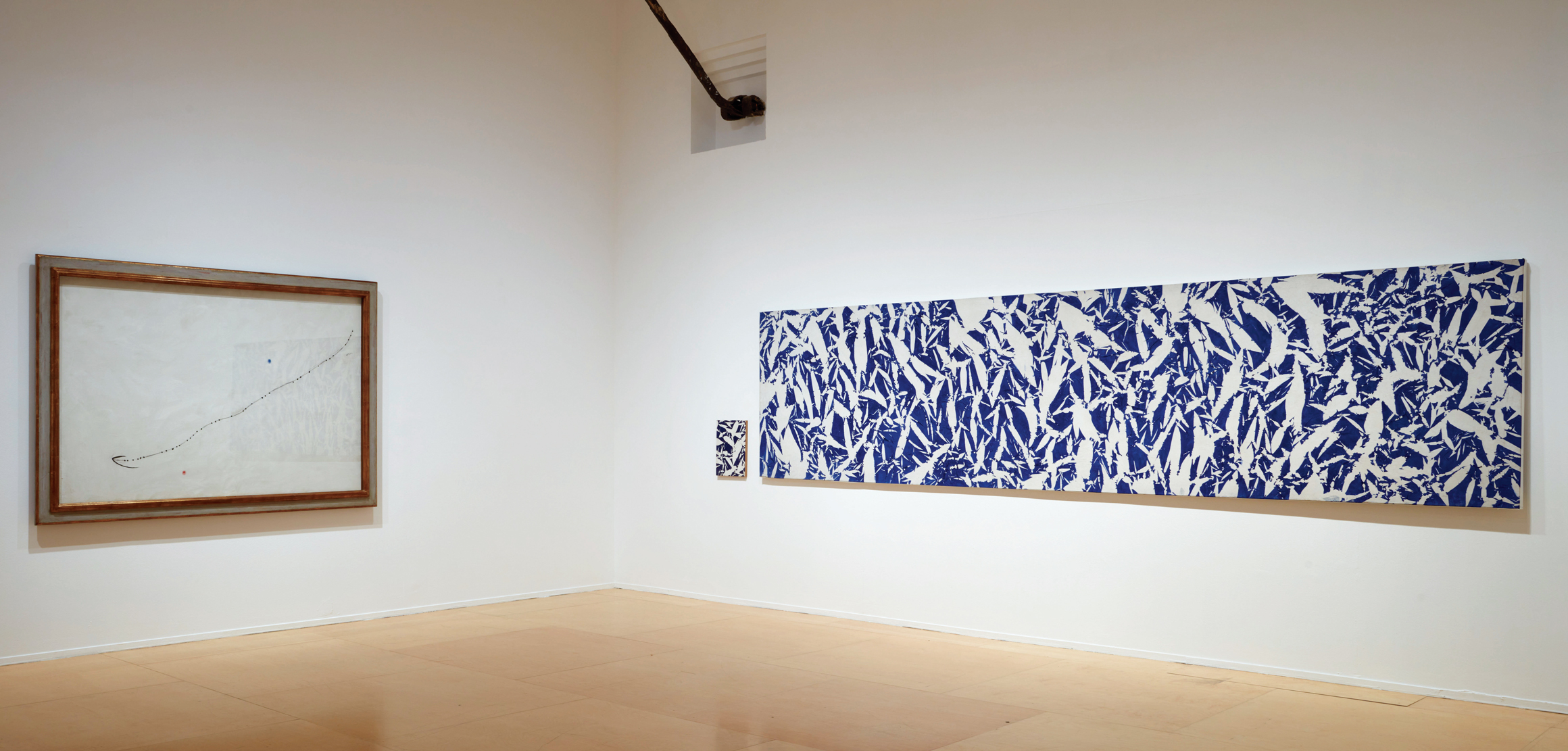 A gallery space with white walls displays a framed minimalist artwork to the left and two abstract blue-and-white patterned pieces on the right.