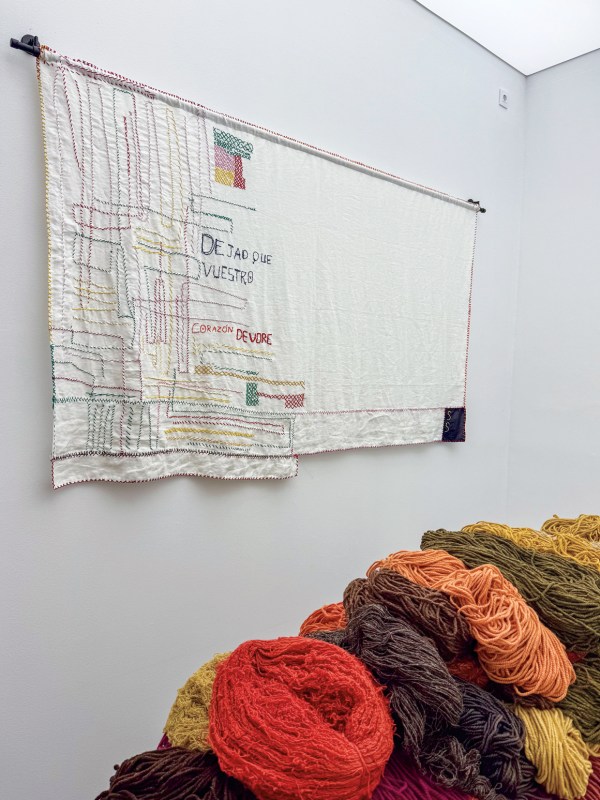 A wall-hung embroidered textile with colorful stitched patterns and text, accompanied by piles of multicolored yarn in shades of red, orange, yellow, and brown.