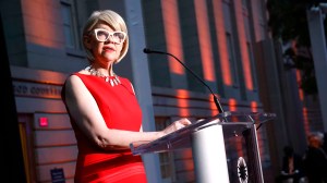 A woman in a red dress is standing behind a podium with a microphone, delivering a speech or presentation. The podium has the Smithsonian National Portrait Gallery logo on it. The background features a building with warm lighting accents.