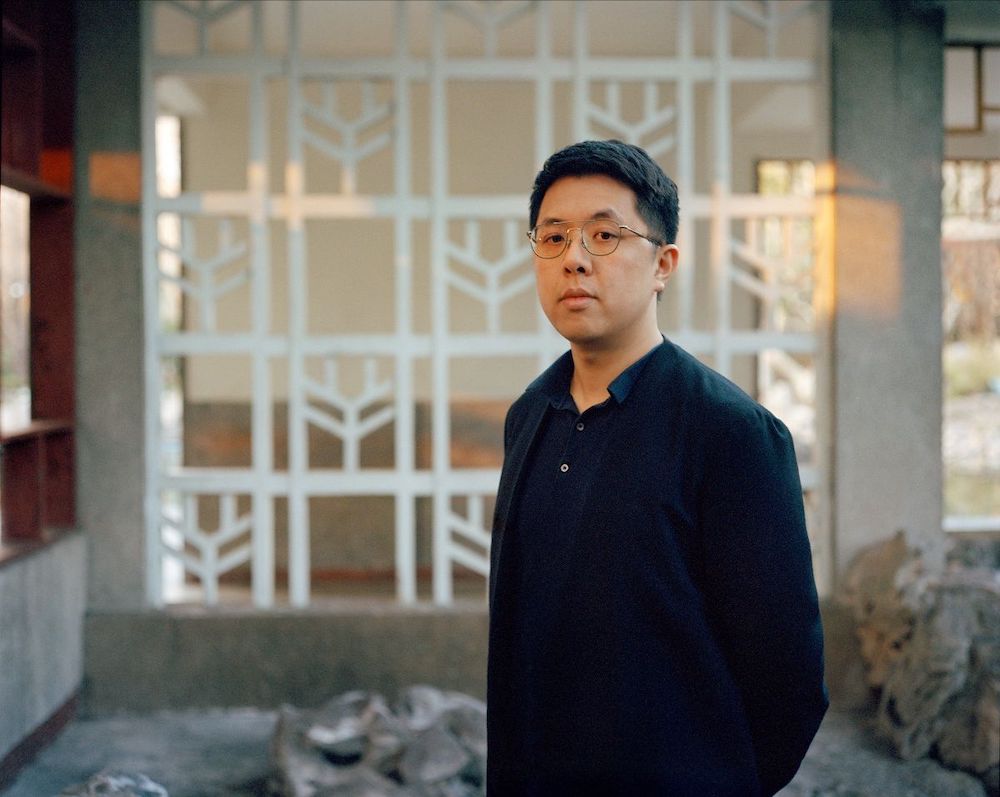 A person wearing a dark shirt is standing in an outdoor space with geometric white structures and rocks in the background. The scene has a calm and natural atmosphere.