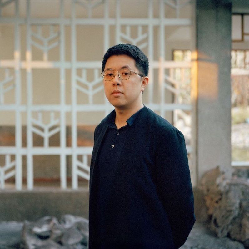 A person wearing a dark shirt is standing in an outdoor space with geometric white structures and rocks in the background. The scene has a calm and natural atmosphere.