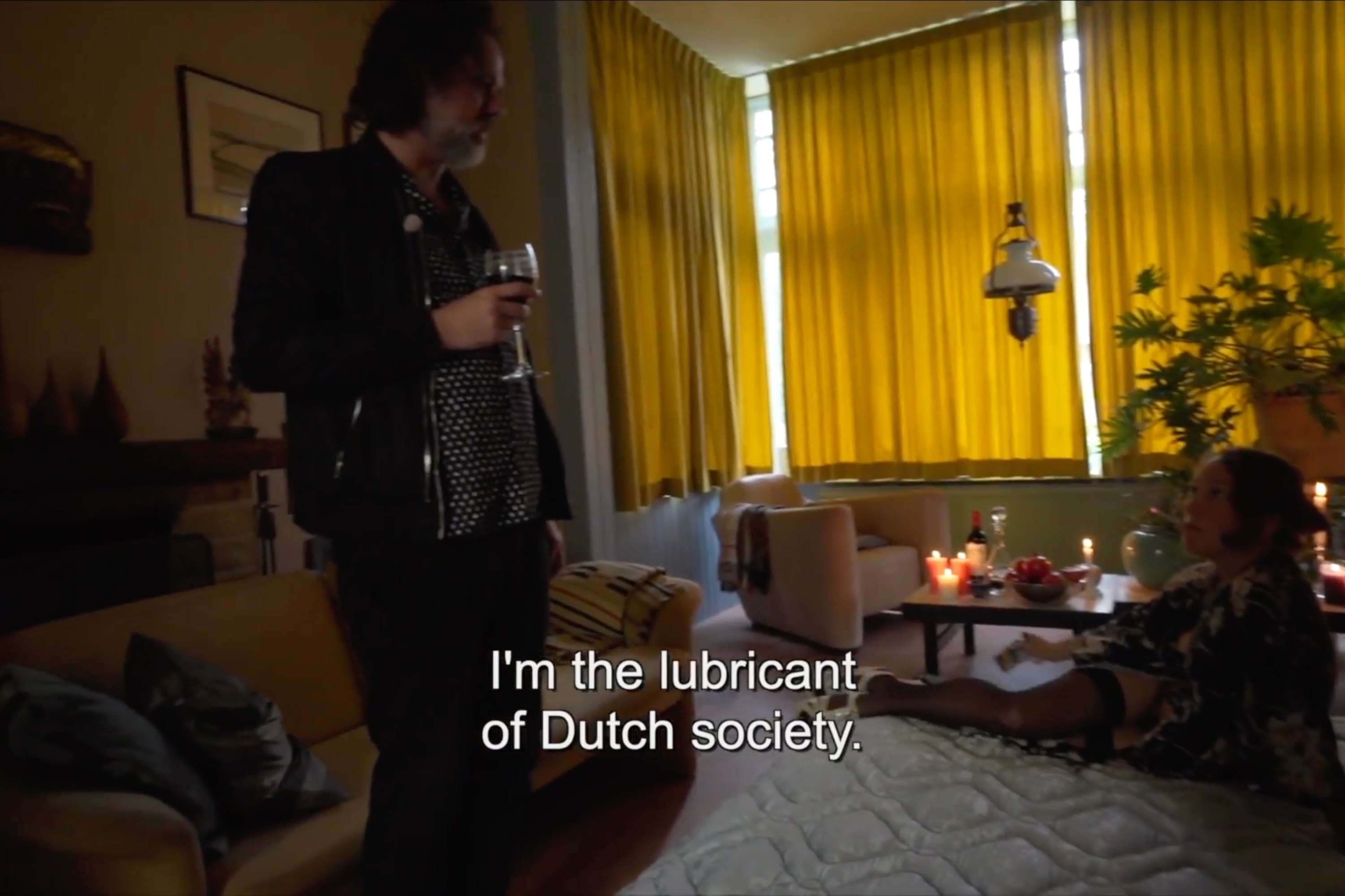 A person stands holding a glass of wine in a dimly lit living room with yellow curtains, candles, plants, and a seated person on a cushioned surface. Subtitles overlay with the words "I'm the lubricant of Dutch society."