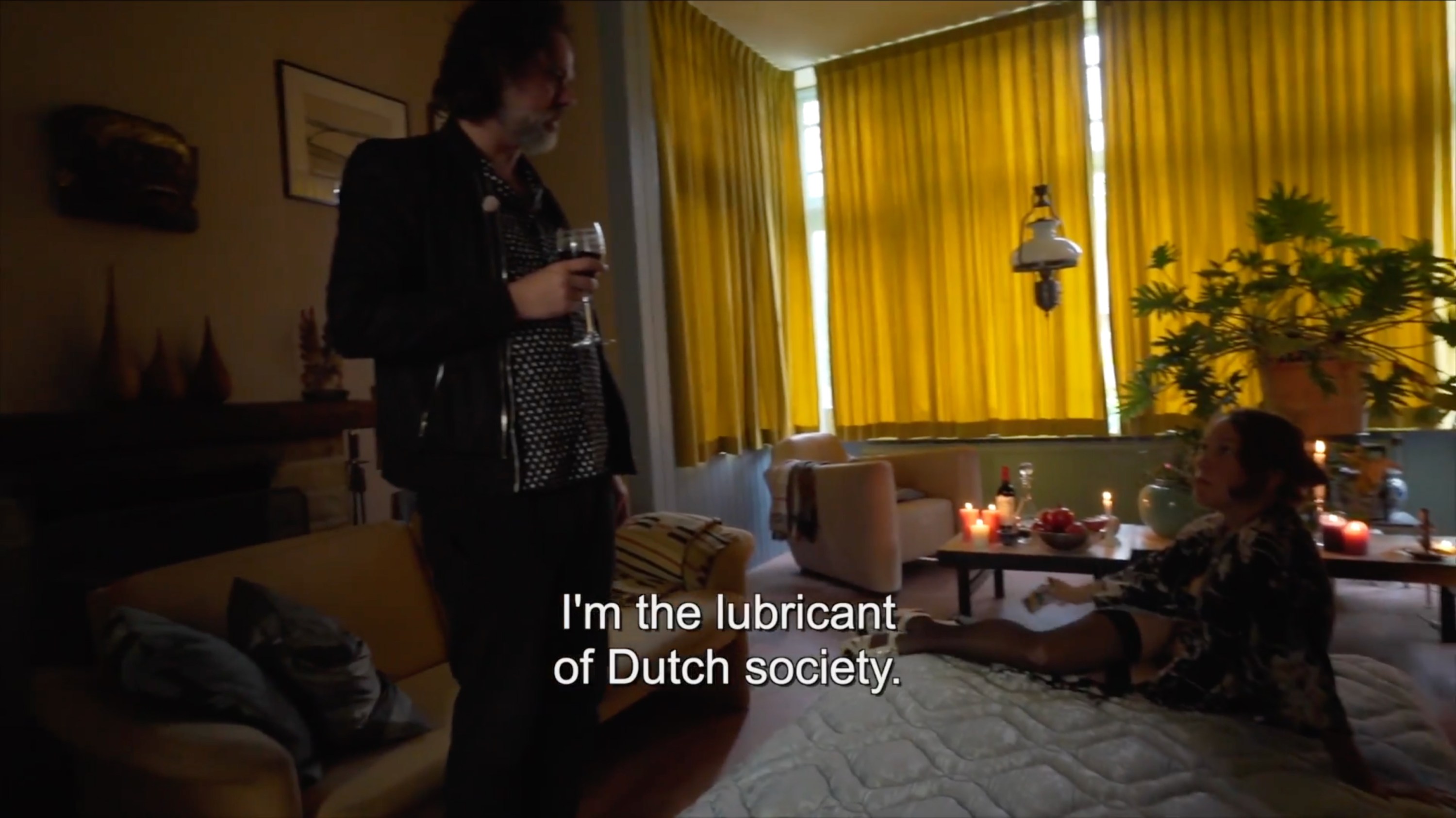 A person stands holding a glass of wine in a dimly lit living room with yellow curtains, candles, plants, and a seated person on a cushioned surface. Subtitles overlay with the words "I'm the lubricant of Dutch society."