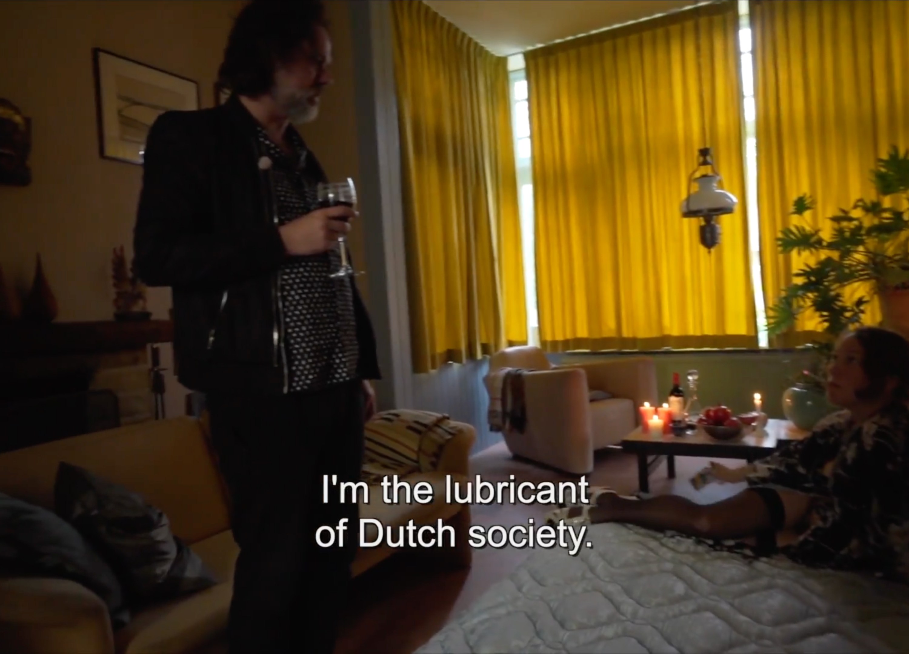 A person stands holding a glass of wine in a dimly lit living room with yellow curtains, candles, plants, and a seated person on a cushioned surface. Subtitles overlay with the words "I'm the lubricant of Dutch society."