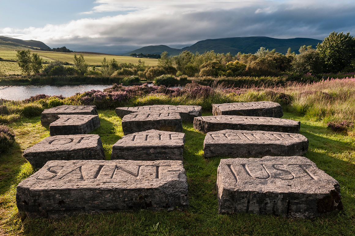 Ian Hamilton Finlay’s Little Sparta, Pentland Hills, Scotland. Large carved stone slabs with inscriptions are arranged on grass near a pond, surrounded by rolling hills, trees, and patches of purple flowers.