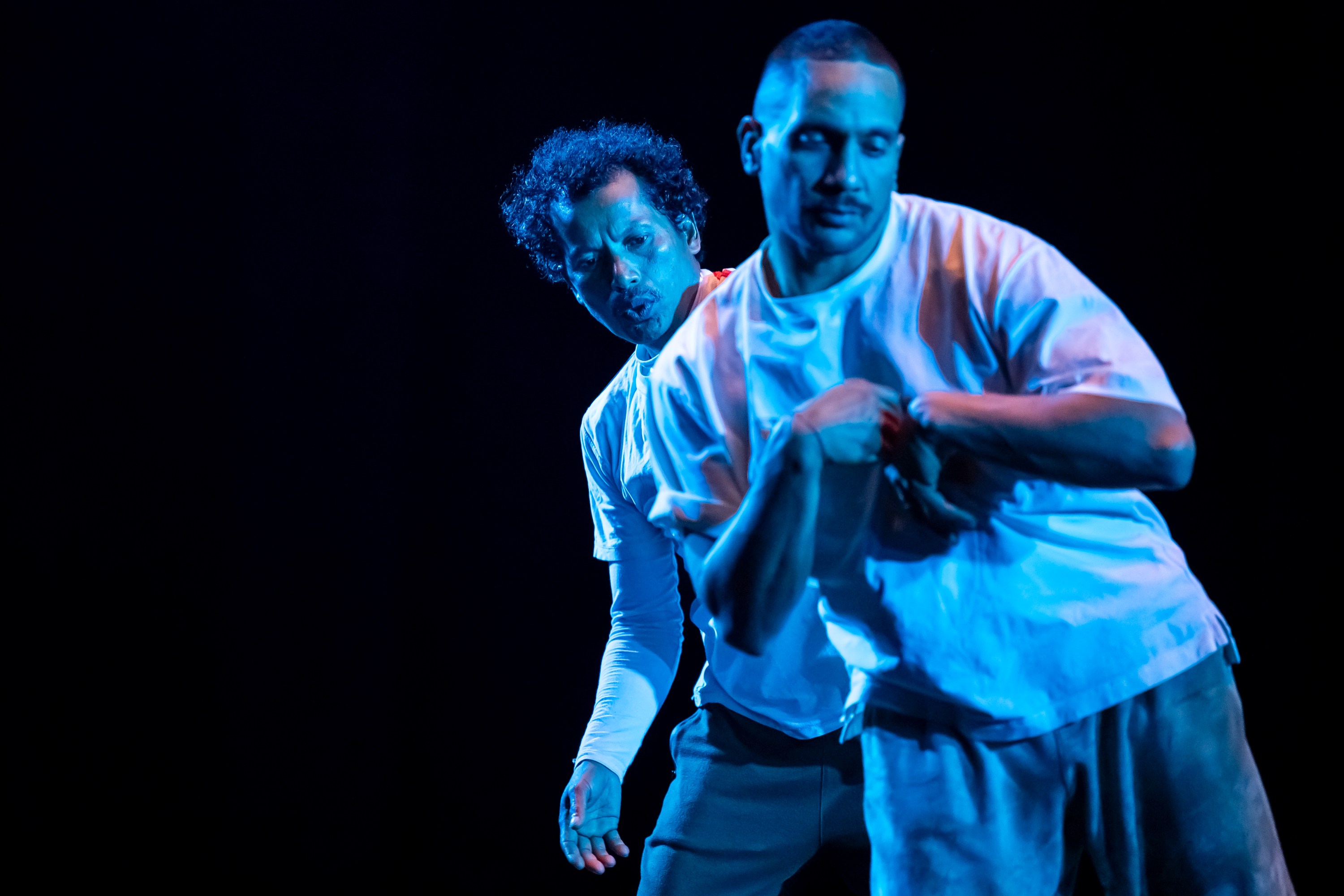Two dance performers in white shirts posed under dramatic blue lighting, one positioned behind the other, with visible arm gestures.