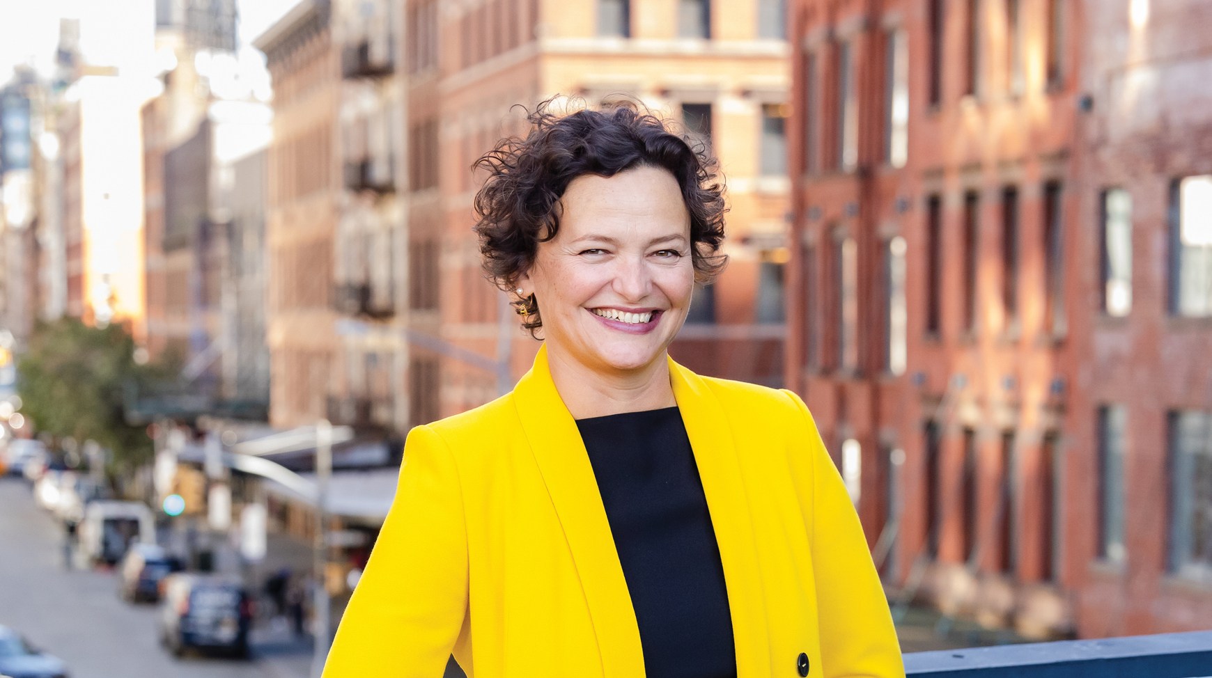 Cecilia Alemani. A person wearing a bright yellow double-breasted coat stands on a railing overlooking a city street with cars, trees, and brick buildings.