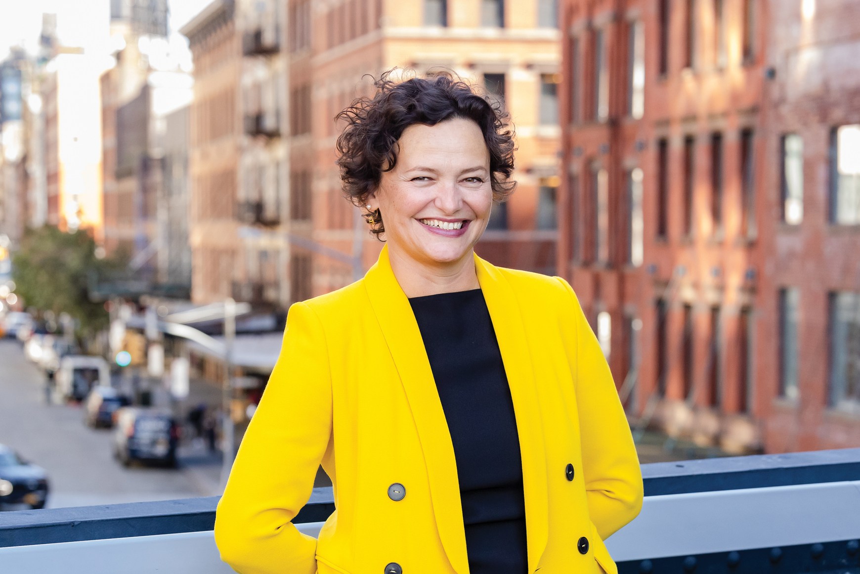Cecilia Alemani. A person wearing a bright yellow double-breasted coat stands on a railing overlooking a city street with cars, trees, and brick buildings.
