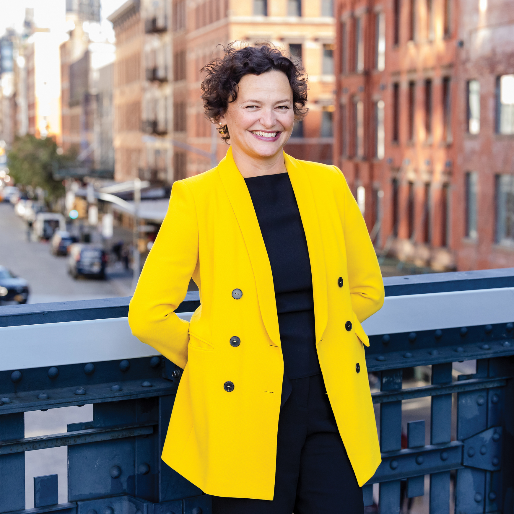 Cecilia Alemani. A person wearing a bright yellow double-breasted coat stands on a railing overlooking a city street with cars, trees, and brick buildings.
