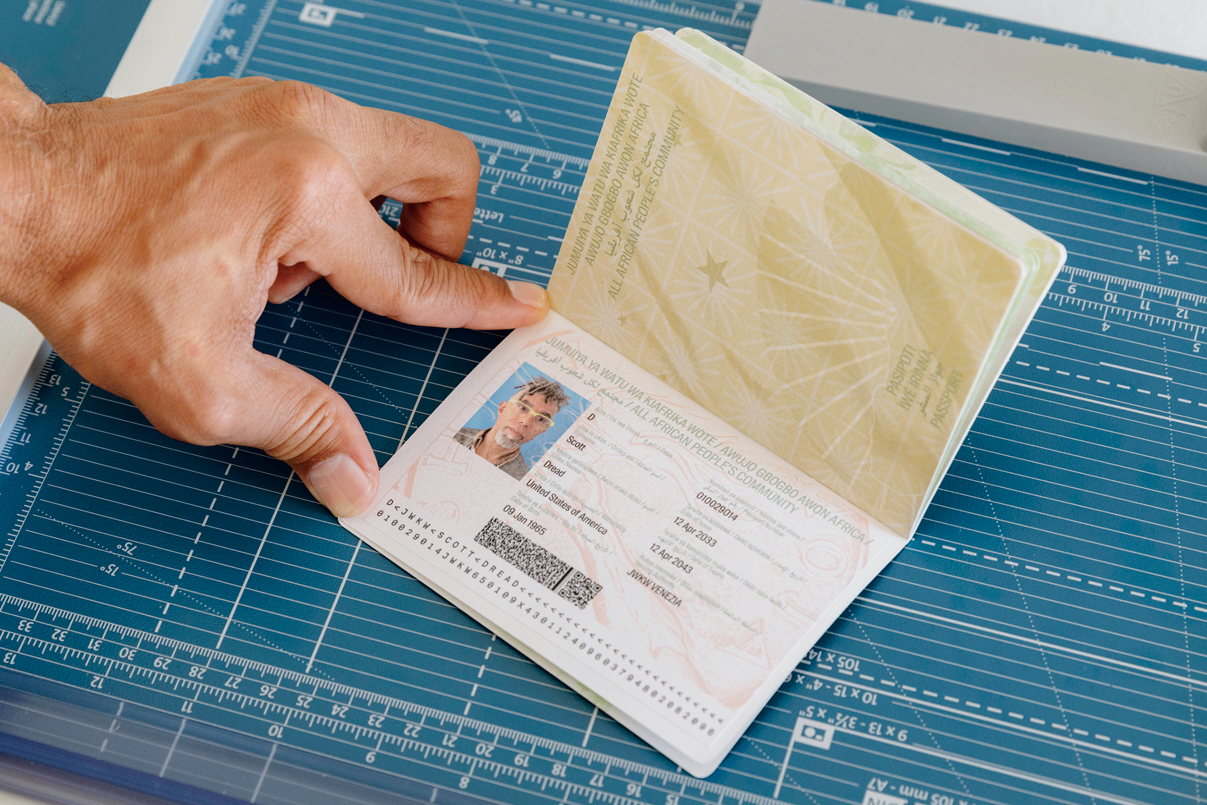 Dread Scott, All African People’s Community Passport. A hand holding an open passport on a blue cutting mat, showing personal details, QR code, and patterned green design on the opposite page.