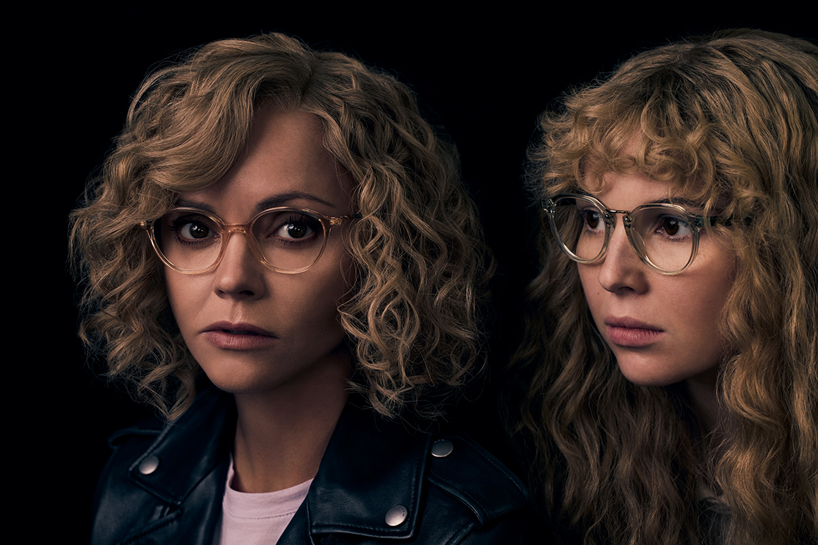 Two individuals with blonde curly hair wearing glasses; one is dressed in a black leather jacket and pink shirt, against a dark background.