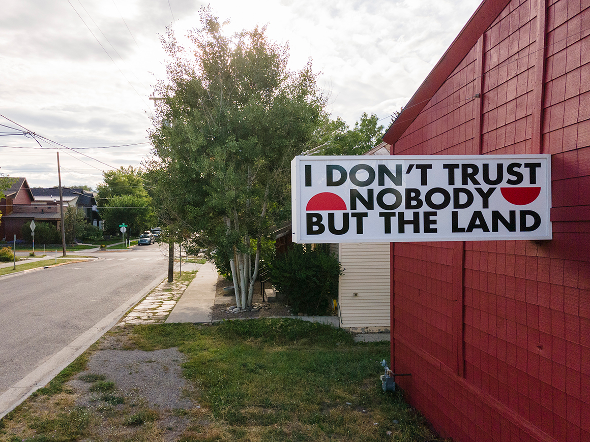 A large banner reading "I DON'T TRUST NOBODY BUT THE LAND" affixed to a red wooden building beside a street in a residential area, with trees and houses visible in the background under a cloudy sky.