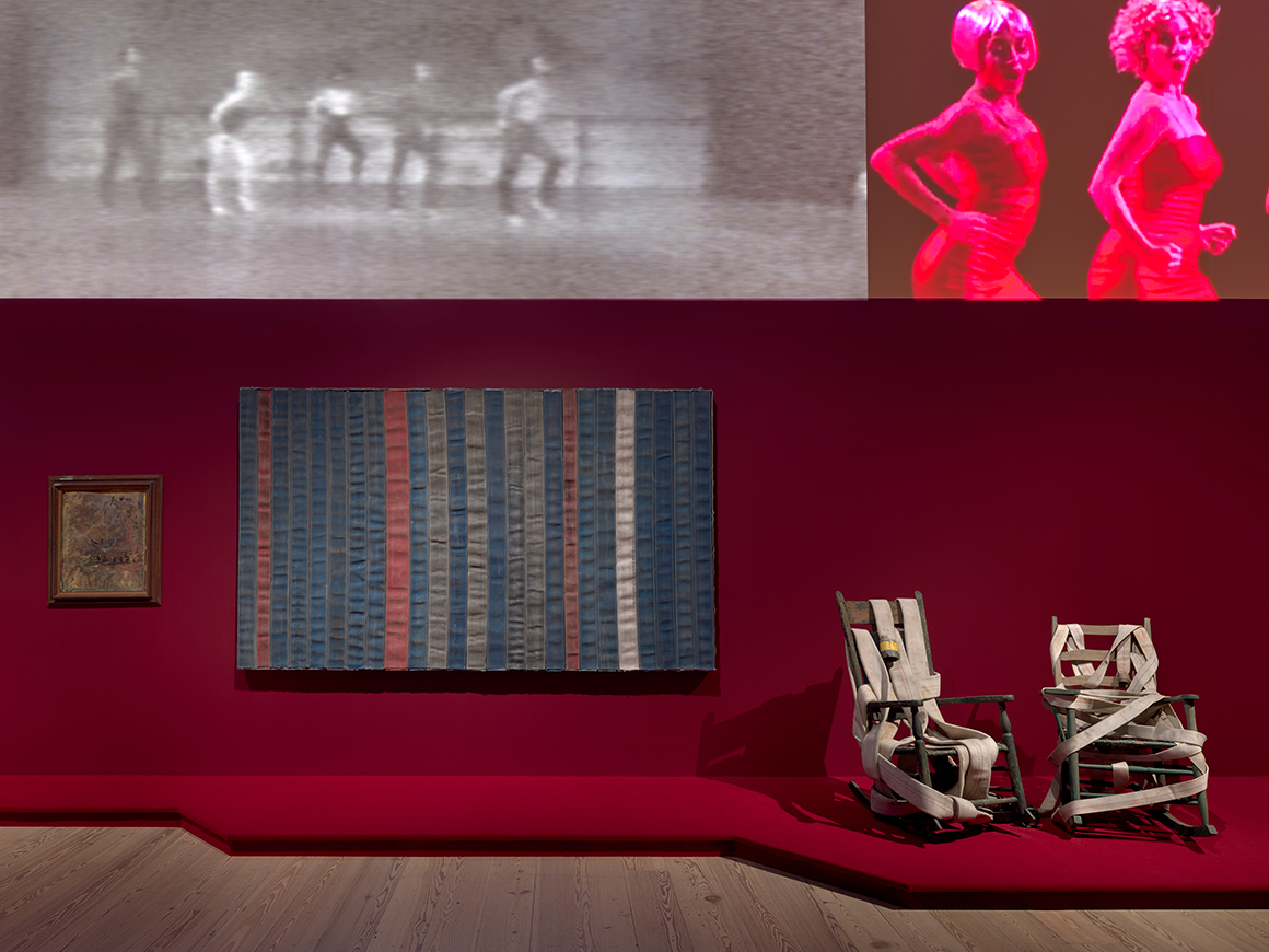 Art gallery with a red wall displaying various artworks: on the left, a small framed traditional painting; in the center, a large rectangular textile artwork with vertical stripes; and on the right, two unconventional white rocking chairs. Above, a digital screen shows two separate projections of people dancing, one in black and white, and the other in color.