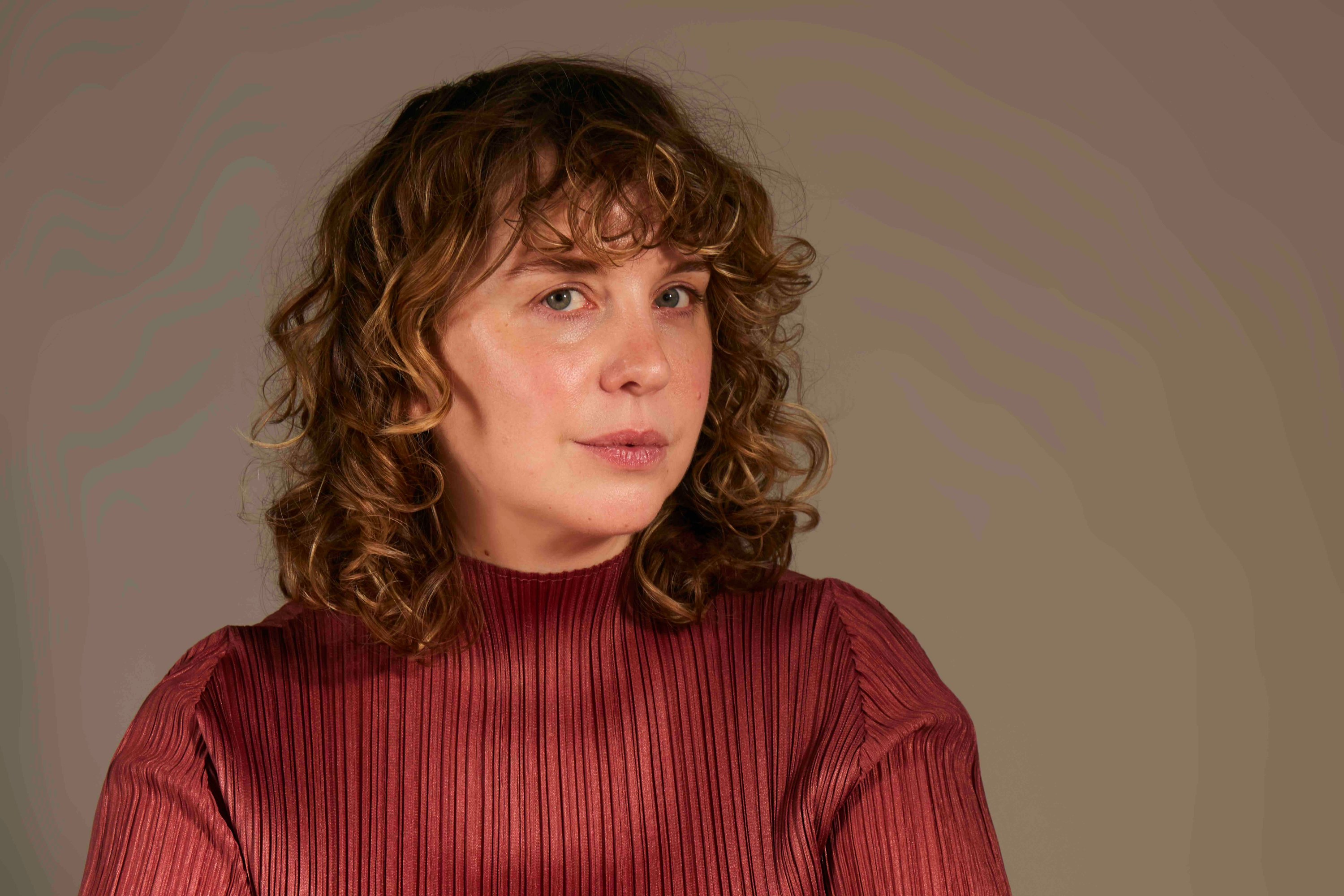 A woman with curly hair wearing a red, textured shirt against a neutral background.
