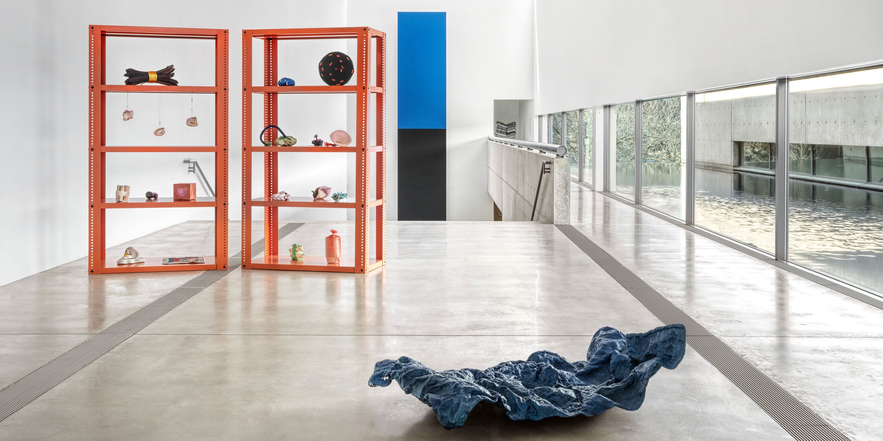 View of “Veronica Ryan: Unruly Objects,” 2025, Pulitzer Arts Foundation, Saint Louis. Two orange shelving units hold assorted sculptures, a crumpled blue fabric-like object rests on the floor, and large windows overlook water and greenery.