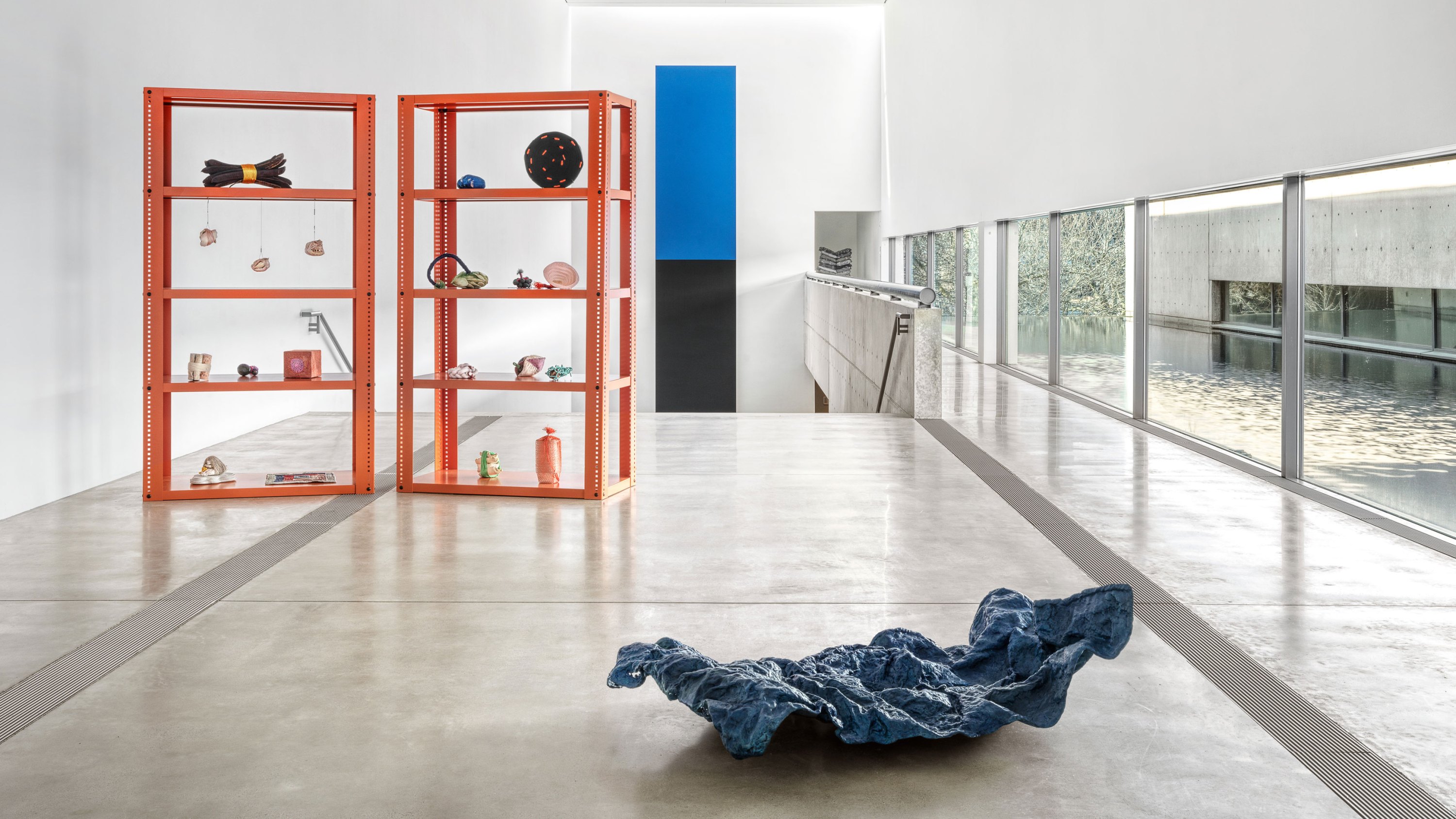 View of “Veronica Ryan: Unruly Objects,” 2025, Pulitzer Arts Foundation, Saint Louis. Two orange shelving units hold assorted sculptures, a crumpled blue fabric-like object rests on the floor, and large windows overlook water and greenery.