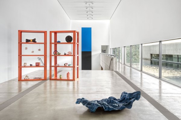 View of “Veronica Ryan: Unruly Objects,” 2025, Pulitzer Arts Foundation, Saint Louis. Two orange shelving units hold assorted sculptures, a crumpled blue fabric-like object rests on the floor, and large windows overlook water and greenery.