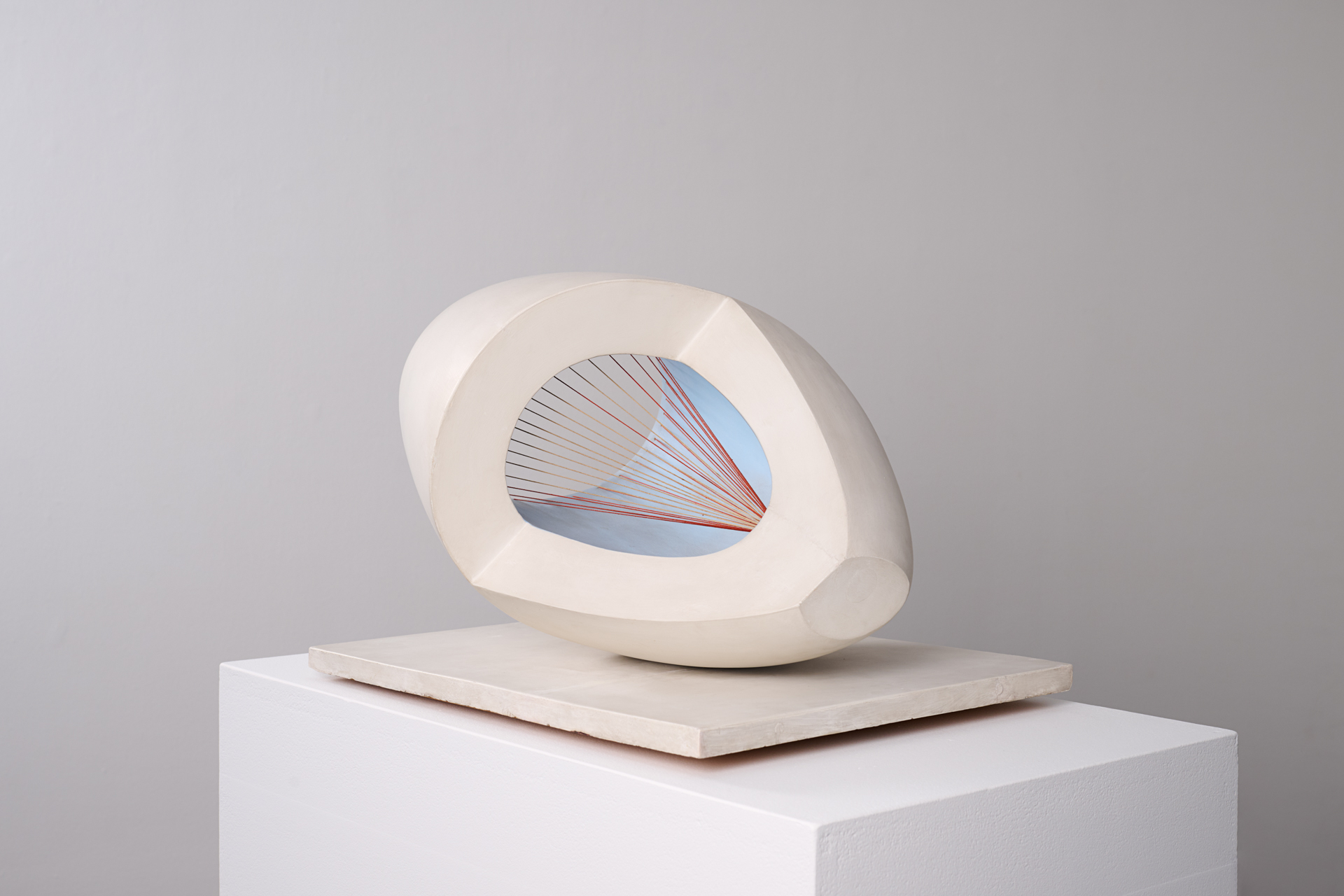 A modern abstract sculpture with a smooth, off-white oval shape featuring a hollow center. Inside, red strings are stretched in a geometric pattern against a light blue background. The sculpture is placed on a square white base.