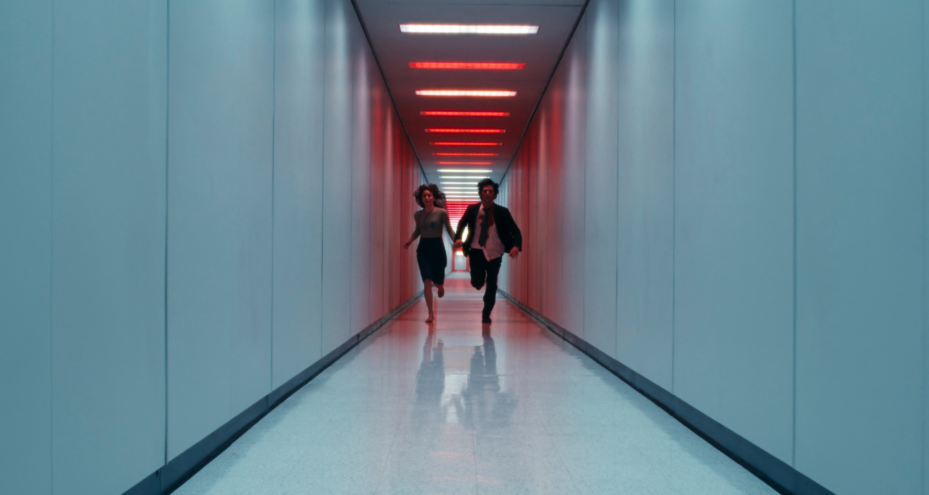 A man and woman are running down a long corridor with light blue walls, a white floor, and red rectangular ceiling lights. Still from the TV show Severance.