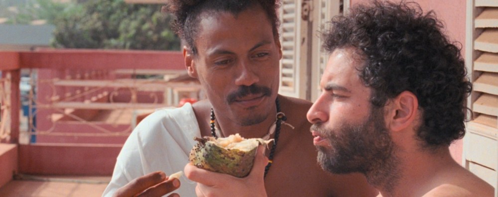 Two people are holding and eating from a halved fruit, with a pink balcony railing, trees, and shutters visible in the background.