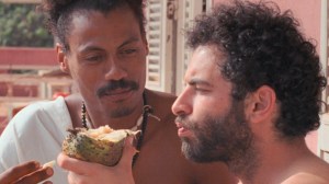 Two people are holding and eating from a halved fruit, with a pink balcony railing, trees, and shutters visible in the background.