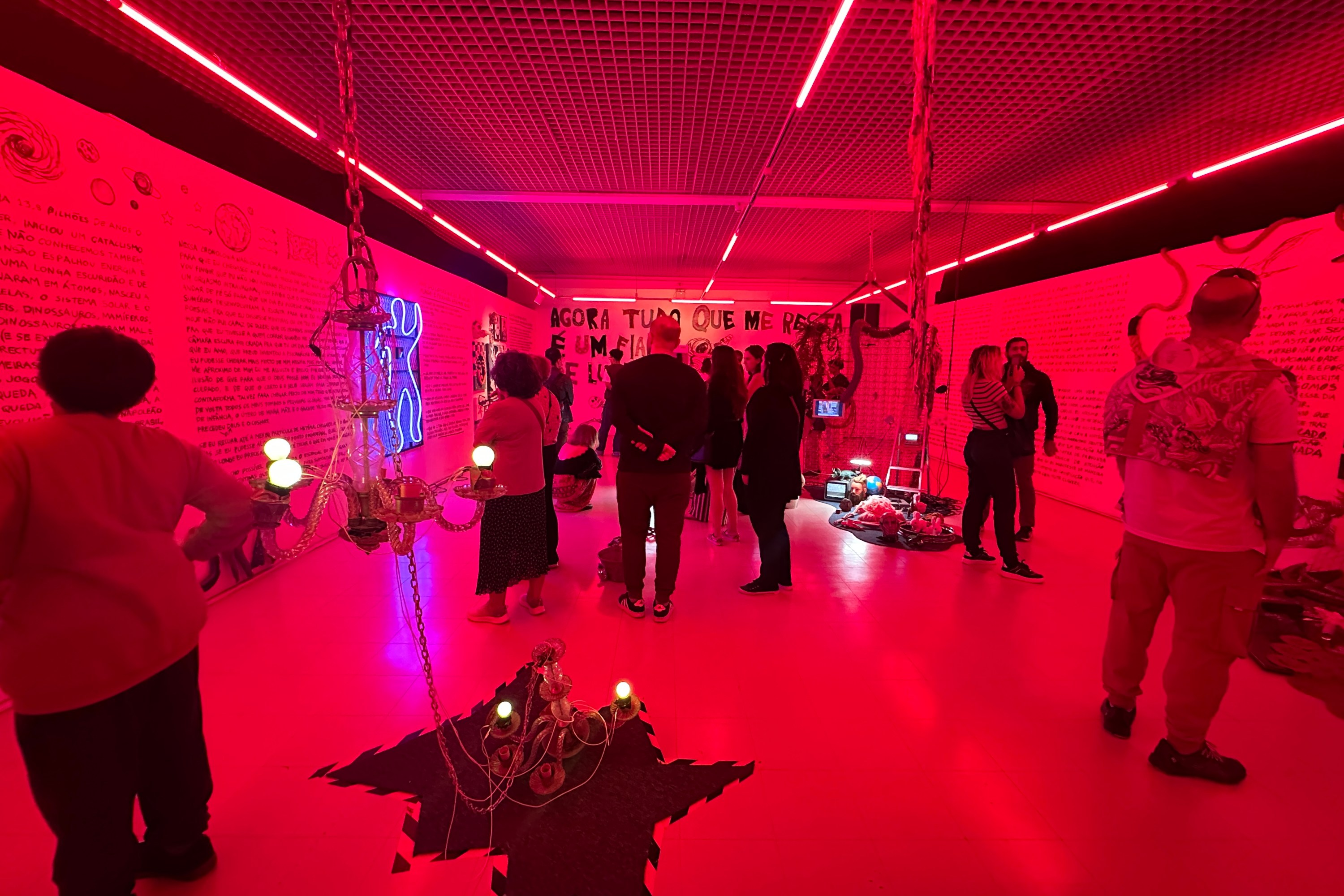 People standing in a red-lit room with handwritten text on walls, neon art, hanging objects, and various installations on the floor and walls.