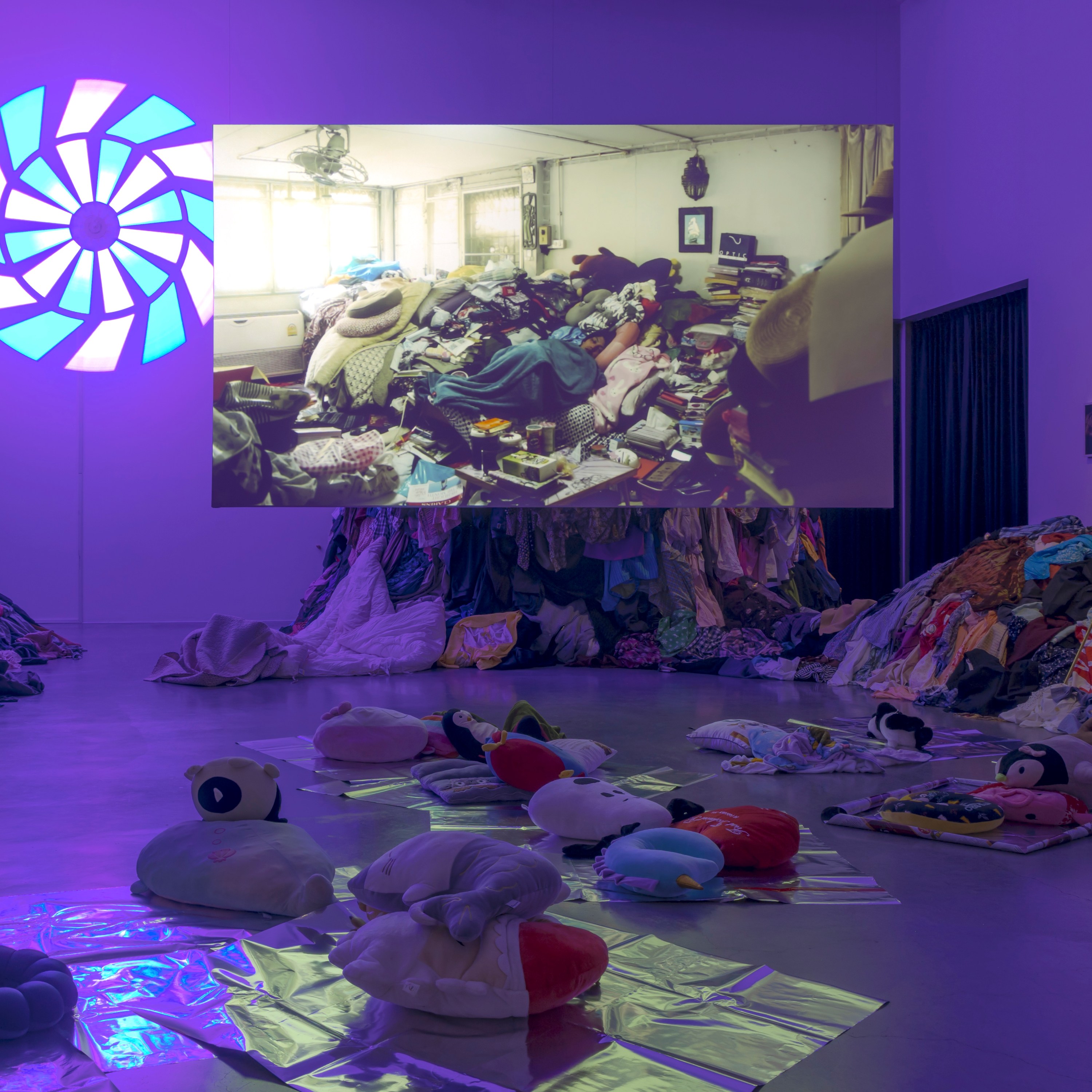 An indoor art exhibit with purple lighting. There are piles of colorful clothes stacked up on the floor and walls. Soft toys, like stuffed animals, are placed on shiny mats around the room. On one side, there's a large circular glowing shape on the wall, resembling a wheel. A projection on the wall shows a cluttered space filled with household items and more clothes. The scene feels unusual and creative.