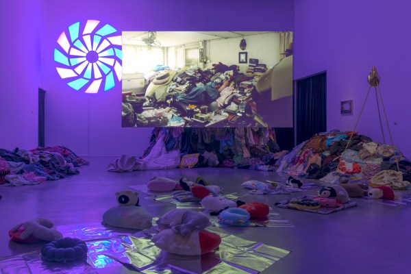 An indoor art exhibit with purple lighting. There are piles of colorful clothes stacked up on the floor and walls. Soft toys, like stuffed animals, are placed on shiny mats around the room. On one side, there's a large circular glowing shape on the wall, resembling a wheel. A projection on the wall shows a cluttered space filled with household items and more clothes. The scene feels unusual and creative.