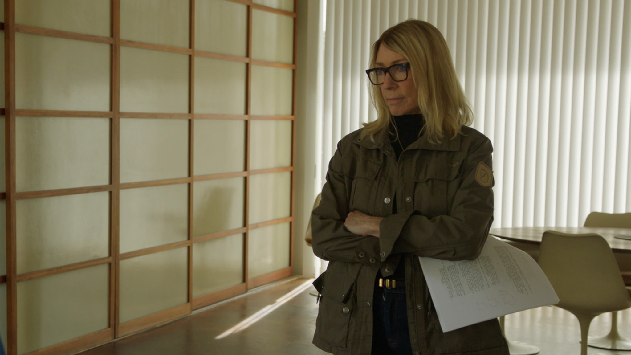 Kim Gordon stands in an empty office with her arms crossed.