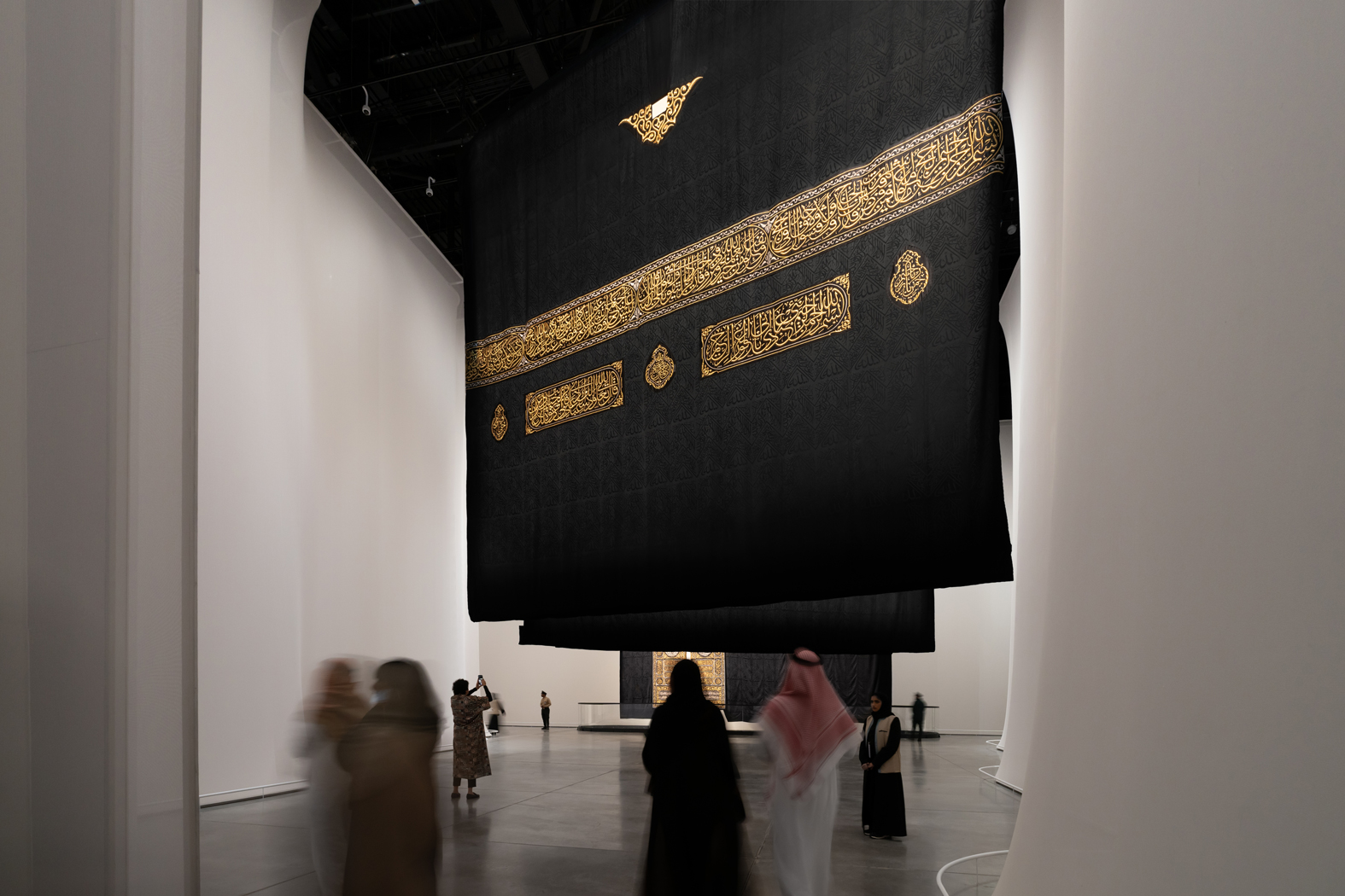 Holy Kaaba kiswah panels. A large black cloth featuring intricate gold calligraphy hangs in a spacious room with people walking, standing, and observing the display.