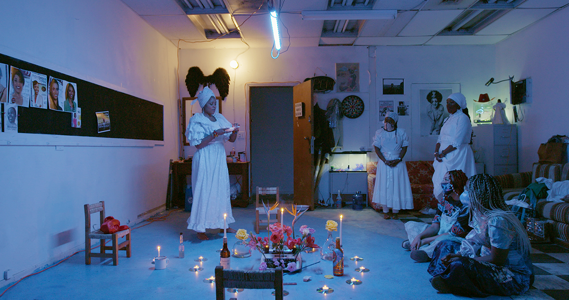 Interior scene of a ritualistic or cultural gathering in a dimly lit room, decorated with numerous candles and a centerpiece with flowers on the floor. Several individuals in traditional white and patterned outfits stand and sit around the space, some observing and others actively participating.