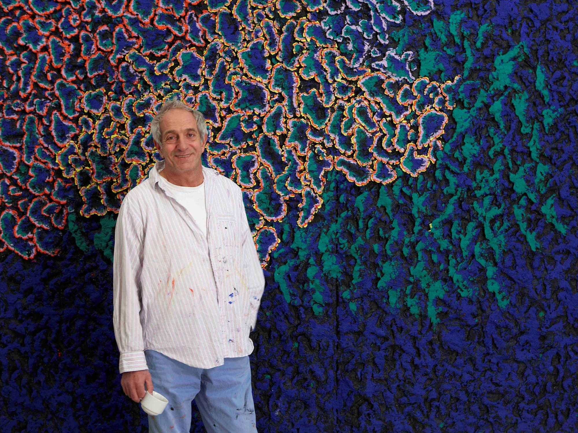 A person wearing a paint-splattered shirt and jeans stands in front of a vibrant abstract artwork featuring intricate, colorful patterns of orange, blue, green, and purple.